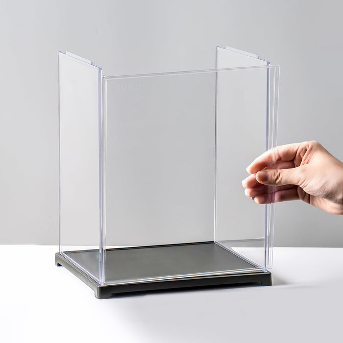 Robotime clear acrylic display case with black base, hand placing it on white surface for figure doll storage